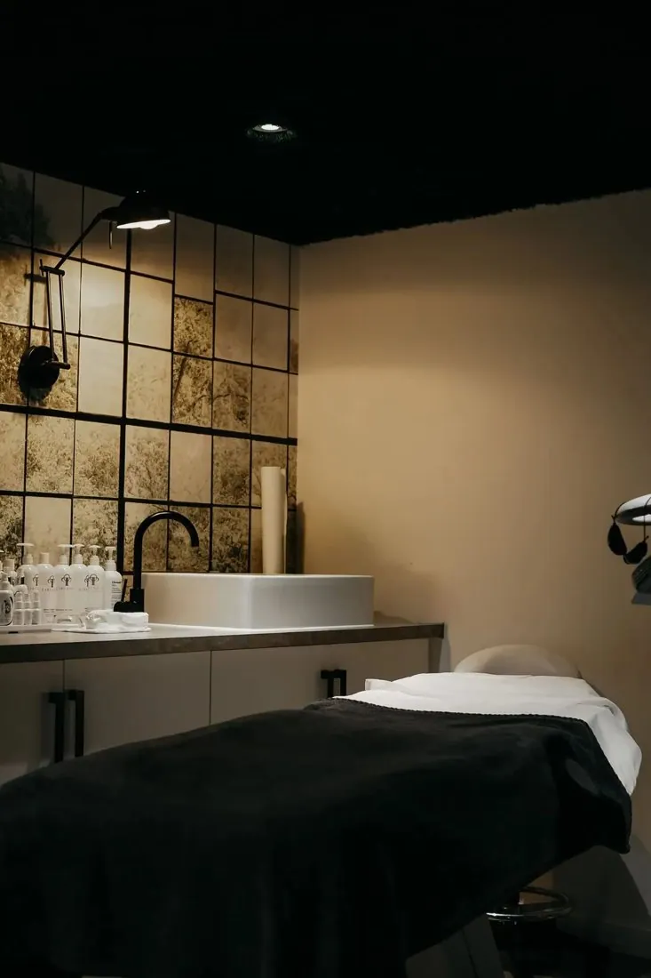 Young LDN treatment room with a dark-covered bed, shelving and sink.